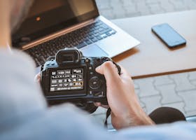 Close-up of hands holding DSLR camera at a workspace with laptop and smartphone.